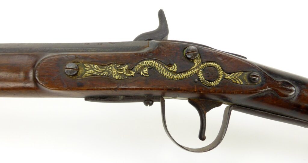 Northwest trade gun neboli severozápadní komerční puška. Detail mosazného odlitku hada nebo draka přišroubovaného na protistranu zámkové desky. Jedná se o typický poznávací znak této zbraně.