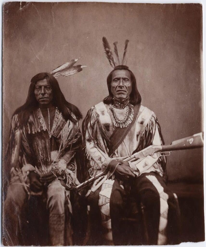 Šílený býk a Tetřívek, bojovníci kmene Atsinů na fotografii Stanley Morrowa z roku 1870. Bojovník vpravo má na kolenou opakovací pušku Henry.