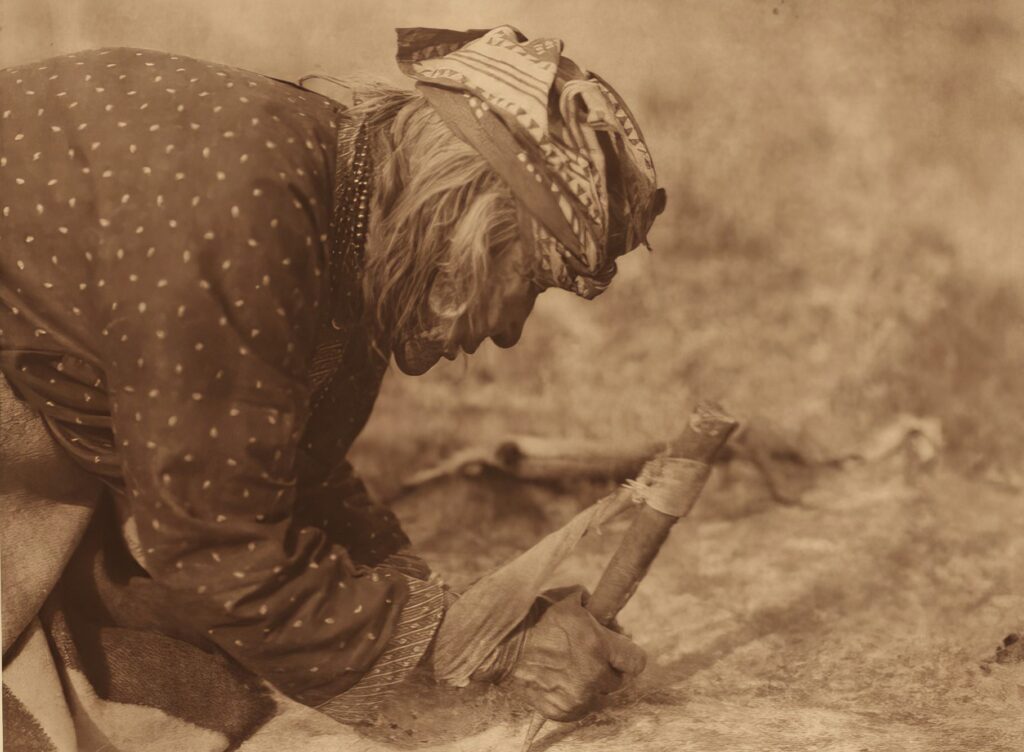 Indiánka na dobové fotografii mízdří kůži pomocí speciální škrabky, které se říká flesher (flešr). Foto E.Curtis.
