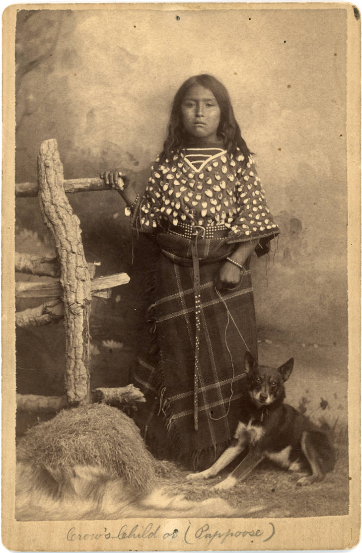 Nedatovaná fotografie dívky z kmene Vran v typických šatech staršího typu s otevřenými a širokými rukávy.