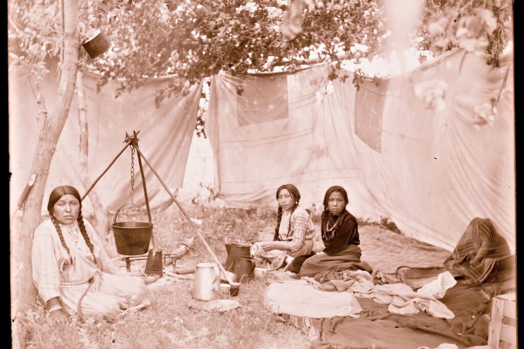 Indiánky kmene Vran vaří venku. Ze starého liningu si vyrobily provizorní zástěnu proti větru a slunci. Dobová fotografie R.Throssela.