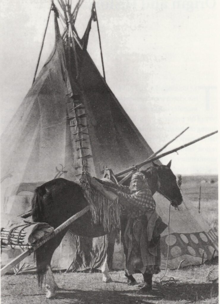 Žena kmene Černonožců na dobové fotografii. Všimněte si parfleše připevněné ke koňskému smyku a tubusu připevněnému k sedlu.