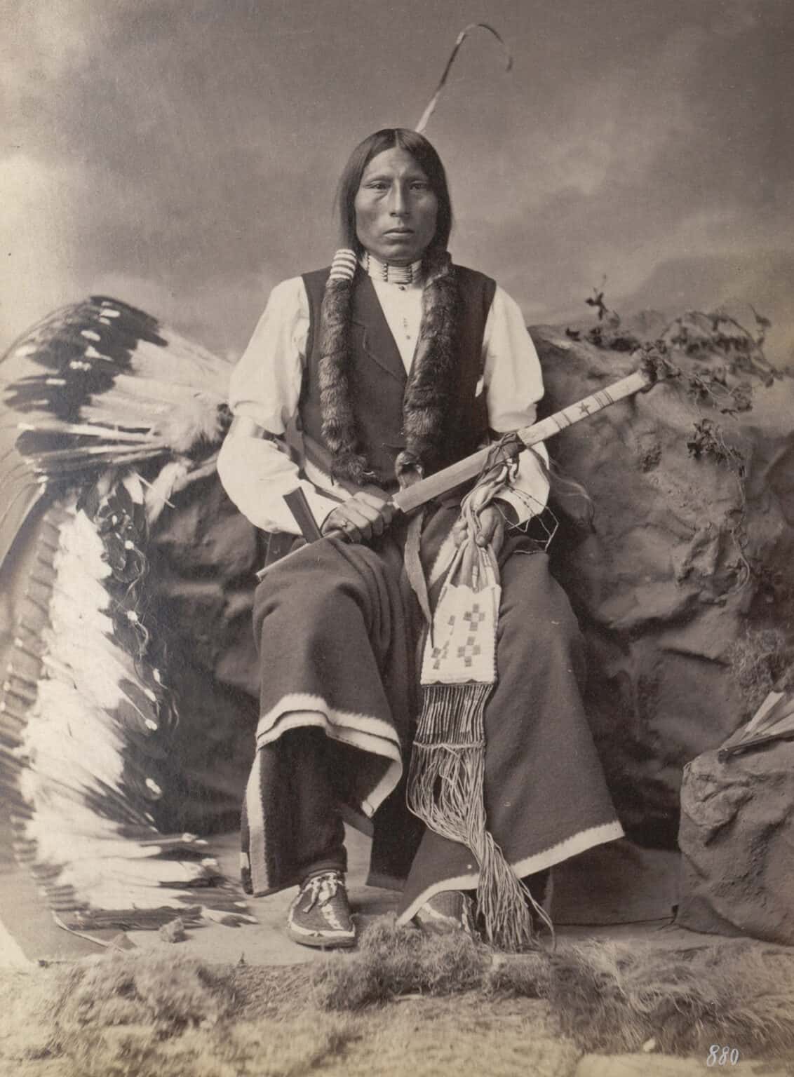 Tři medvědi, oglalský Lakota na fotografii z roku 1877. Jeho legíny jsou vyrobeny z vlněné látky s bílým neobarveným okrajem, stejně jako jako jeho přikrývka na kolenou.